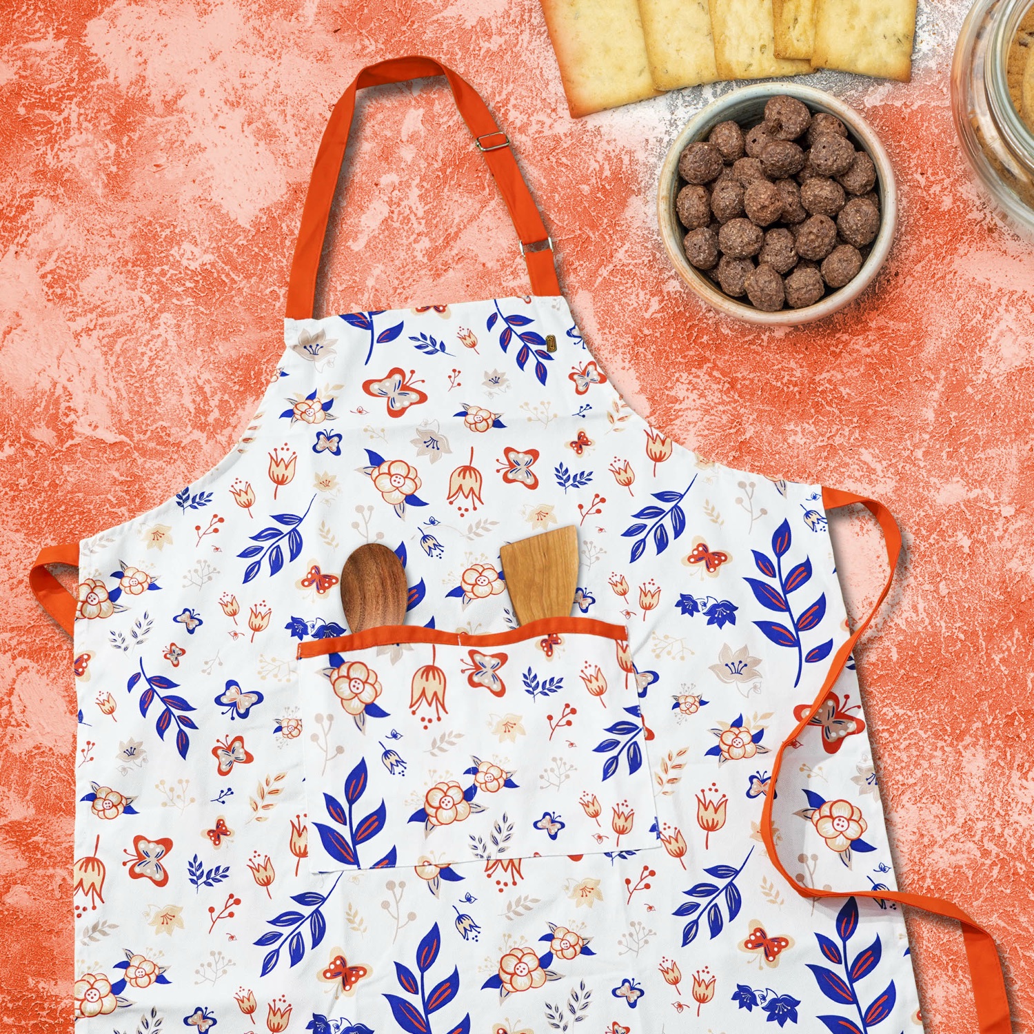 Floral apron with orange ties and double pockets laid flat on a coral surface